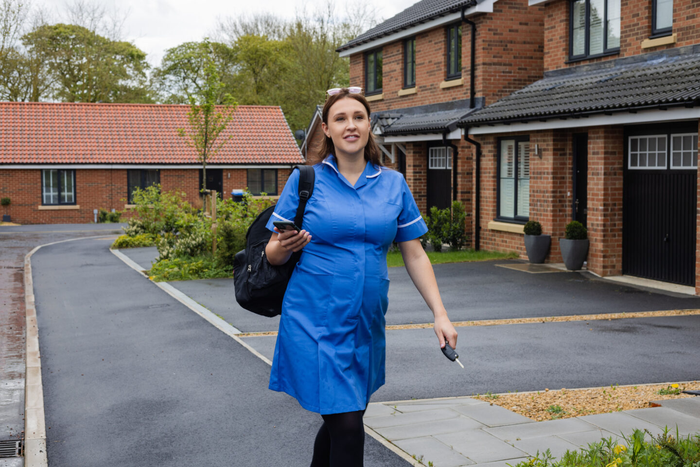 A shot of a young pregnant female walking to her car outside her home in Sedgefield, North East England. She is wearing a nurse uniform ready for work, carrying a backpack over one shoulder and holding her car keys in one hand,  mobile phone in the other. She is looking ahead with a content look on her face.