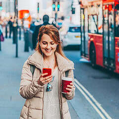 Woman using her phone on a busy London street with a red bus in the background.