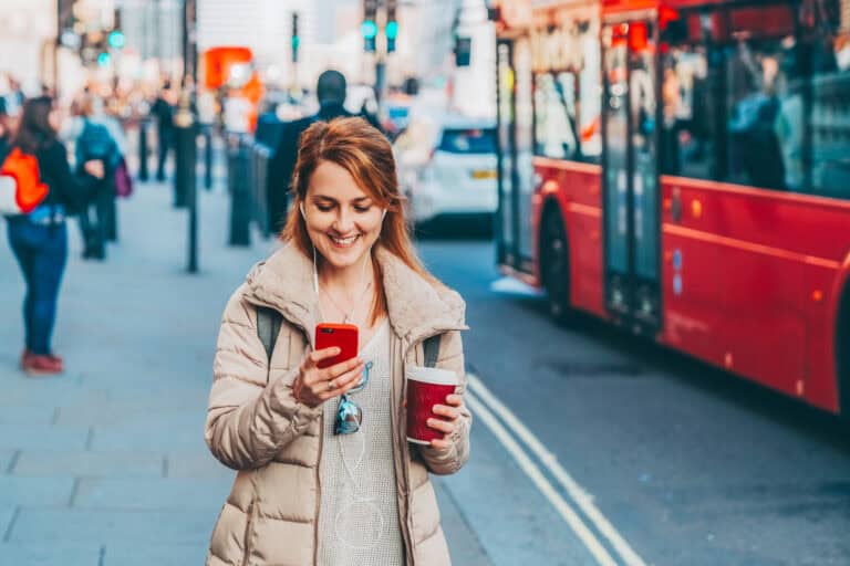 Woman using her phone on a busy London street with a red bus in the background.