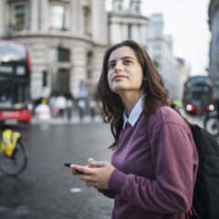 A woman holding a phone stands on a London street, looking thoughtfully into the distance. A red double-decker bus and a cyclist pass behind her.