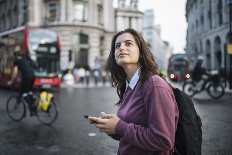 A woman holding a phone stands on a London street, looking thoughtfully into the distance. A red double-decker bus and a cyclist pass behind her.