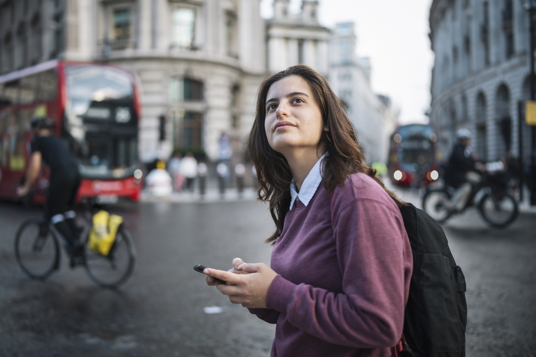 A woman holding a phone stands on a London street, looking thoughtfully into the distance. A red double-decker bus and a cyclist pass behind her.