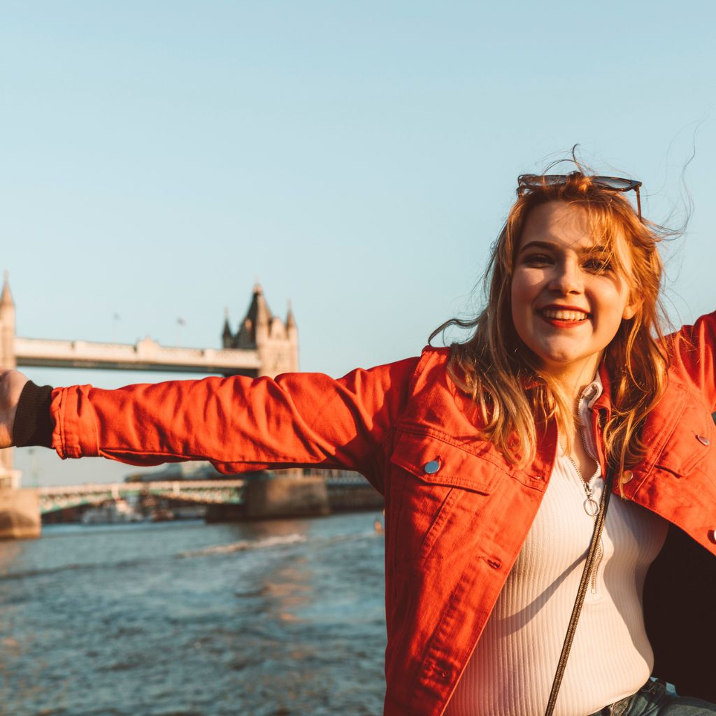 Woman in a red jacket standing by the Thames in front of Tower Bridge with her arms spread out wide